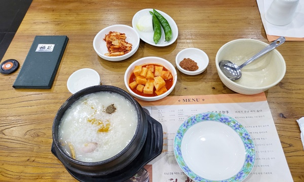 테이블에 삼계탕과 깍두기, 김치, 고추와 양파, 쌈장, 접시, 국자가 올려져있는 사진입니다.