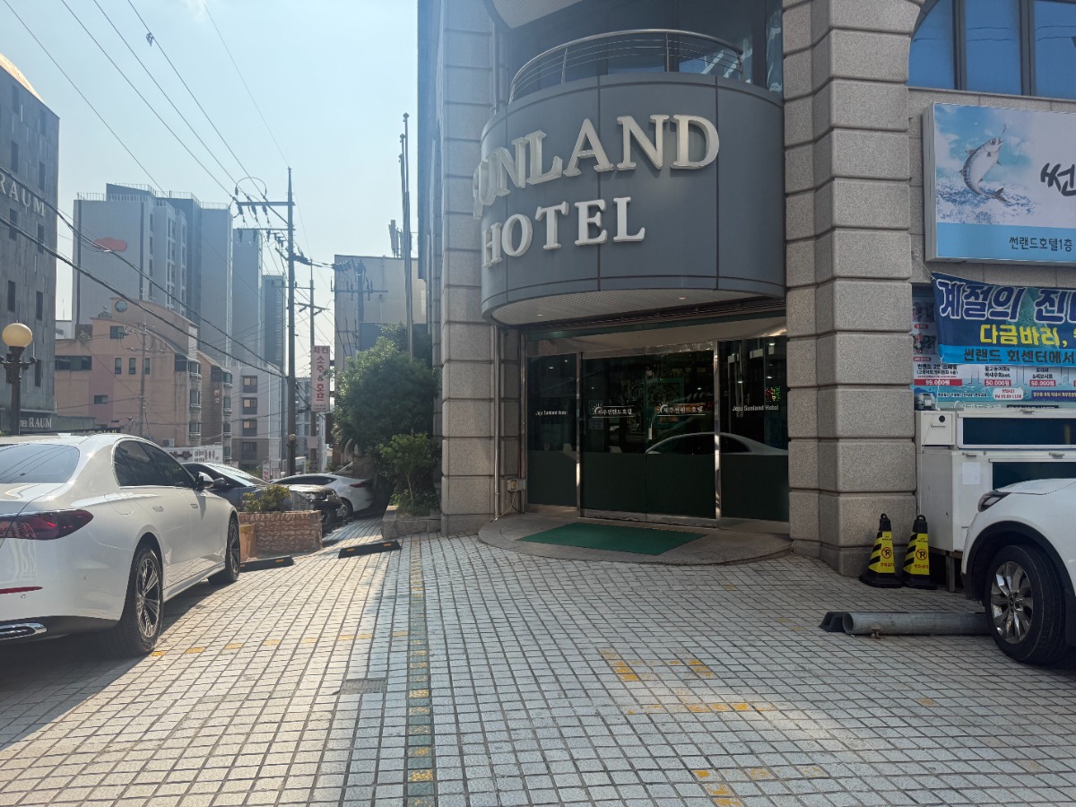 사진에는 SUNLAND HOTEL이라고 적힌 건물의 주출입구가 보이고 있습니다. 출입구는 유리문으로 되어 있으며, 건물 앞 바닥은 회색 계열의 타일로 포장되어 있습니다. 출입구 앞에는 초록색 발판이 놓여 있고, 주출입구 앞 도로에는 흰색 승용차와 SUV 차량이 주차되어 있으며, 인도와 주차 공간이 함께 배치된 구조입니다. 주변에는 고층 건물들이 보이고 전봇대와 전선이 설치되어 있어 도심 속 호텔 입구임을 알 수 있습니다.